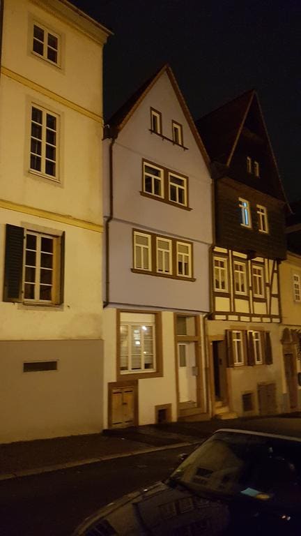 A row of historic buildings is featured at night, highlighting their unique architectural styles. The center building is painted in a soft blue hue, with symmetrical windows and wooden accents. Warm light glows from one window, casting a soft illumination on the façade.