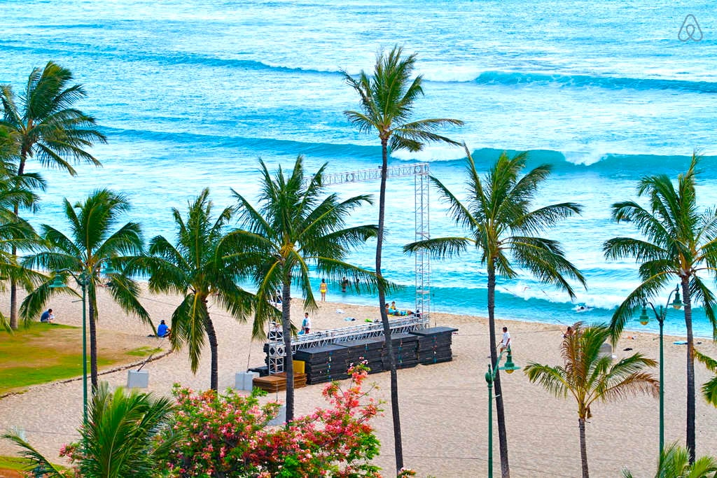 A vibrant beach scene is captured, featuring gentle ocean waves and lush palm trees swaying in the breeze. A stage is set up on the sandy beach, with visitors enjoying the surroundings. The colorful foliage adds to the lively atmosphere.