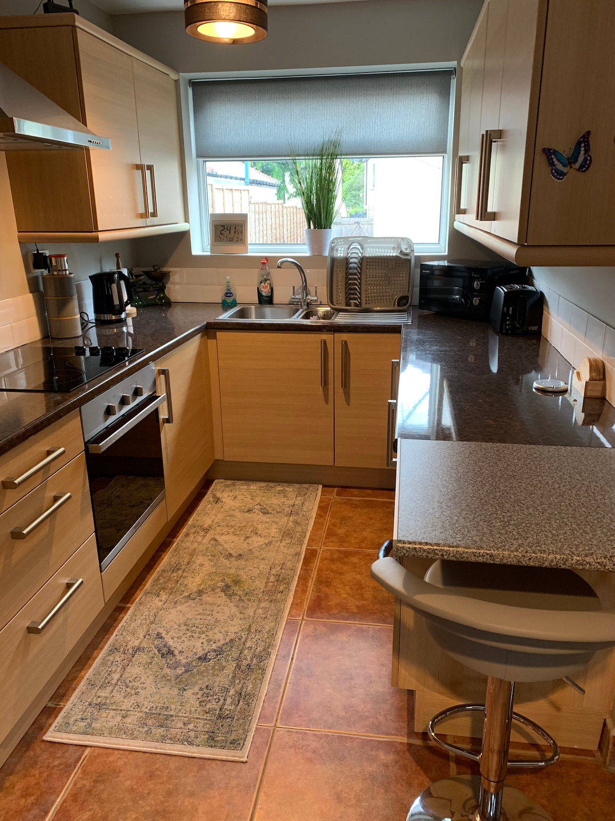 A well-equipped kitchen features wood cabinetry and granite countertops. A sink and stove are positioned adjacent to a refrigerator. A small bar-style table with a stool provides a dining space. Natural light filters in through a window adorned with a roller shade.