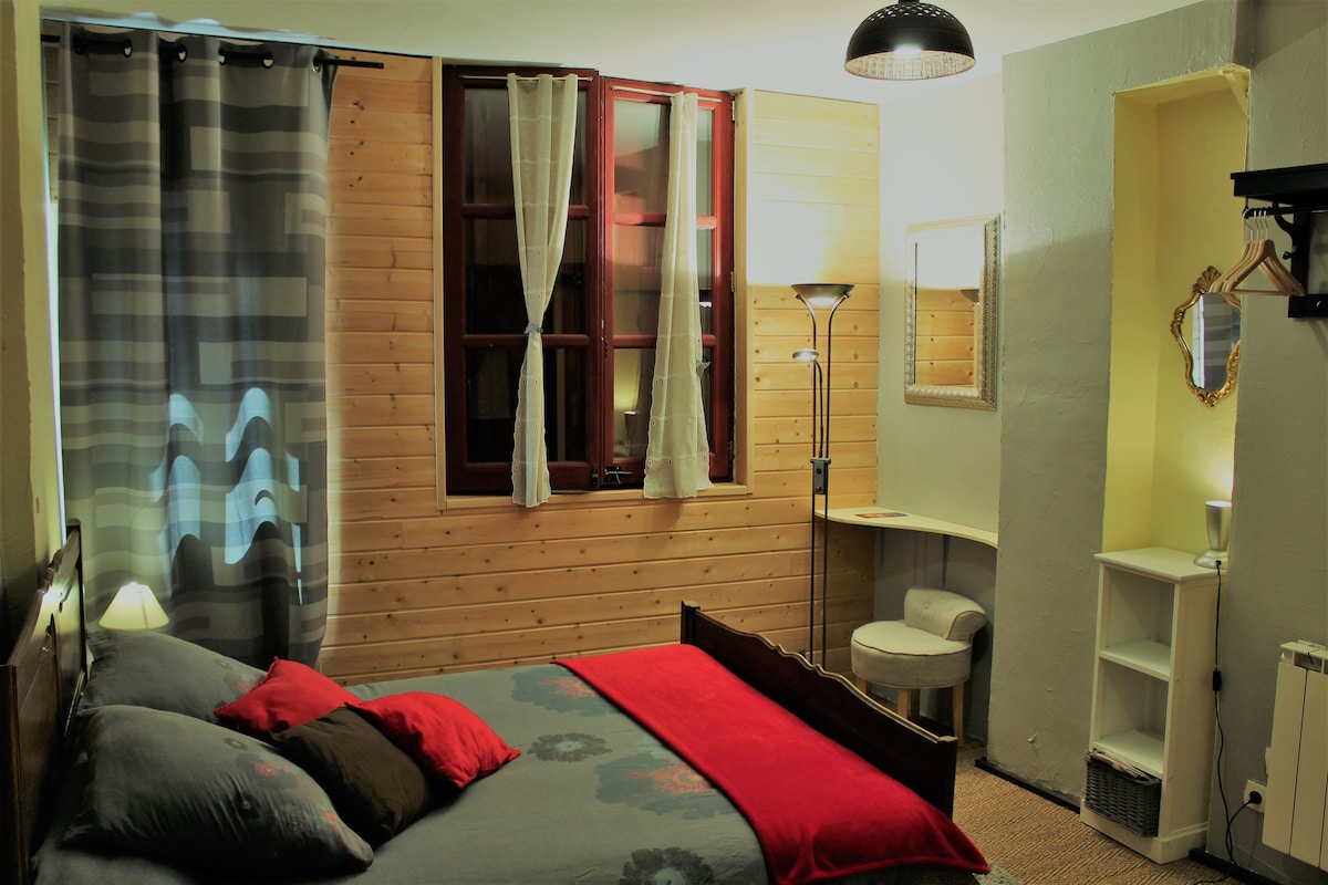 A cozy bedroom features a double bed with a red throw blanket. Natural light enters through window curtains, complementing the wooden paneling on the walls. A small desk and chair, along with a lamp, are positioned beside the bed. A neat shelf is visible in the corner.