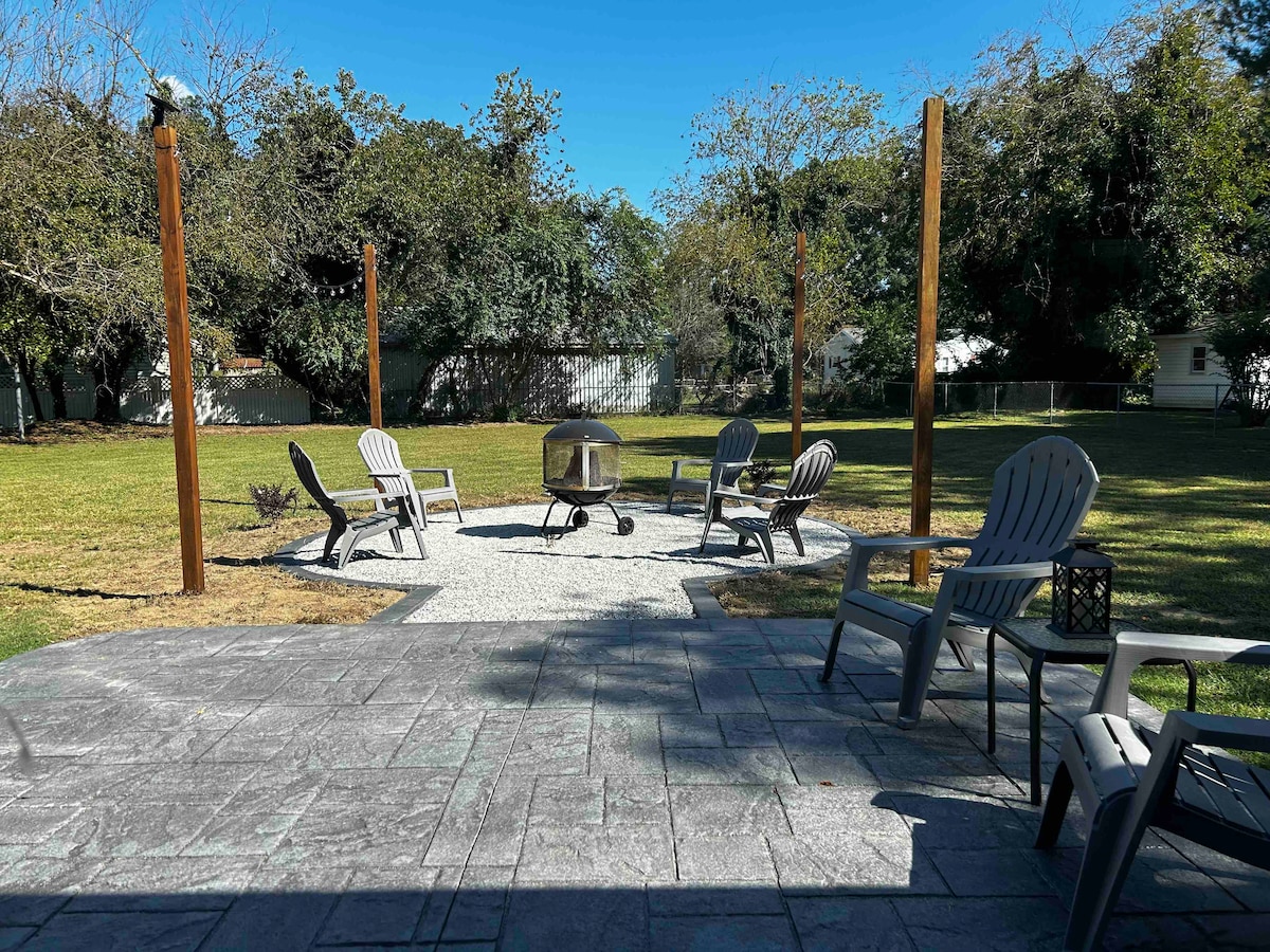 An outdoor patio area features a circular firepit surrounded by six comfortable chairs. The space is bordered by lush green grass and trees, creating a serene setting for gatherings.