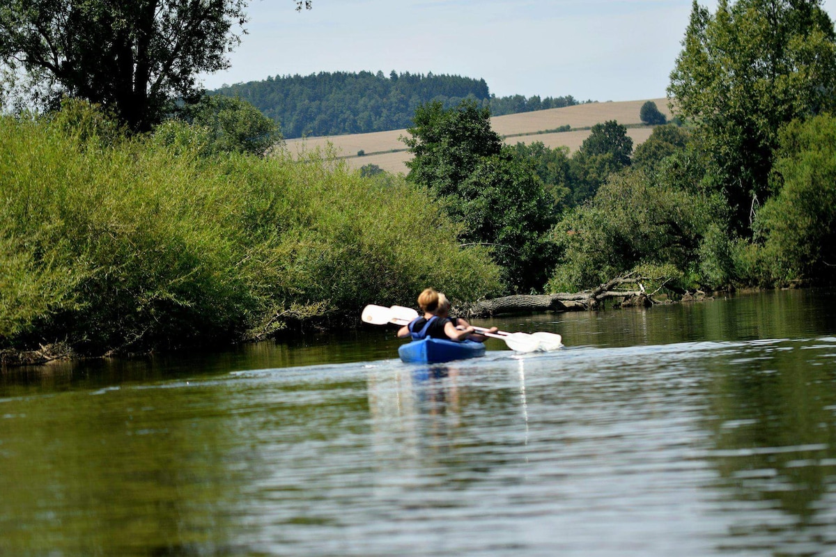 A person is seen kayaking on a calm river, surrounded by lush greenery and trees along the shoreline. The water reflects the blue sky and distant hills, creating a serene atmosphere. Paddles are actively engaged, propelling the kayak forward.