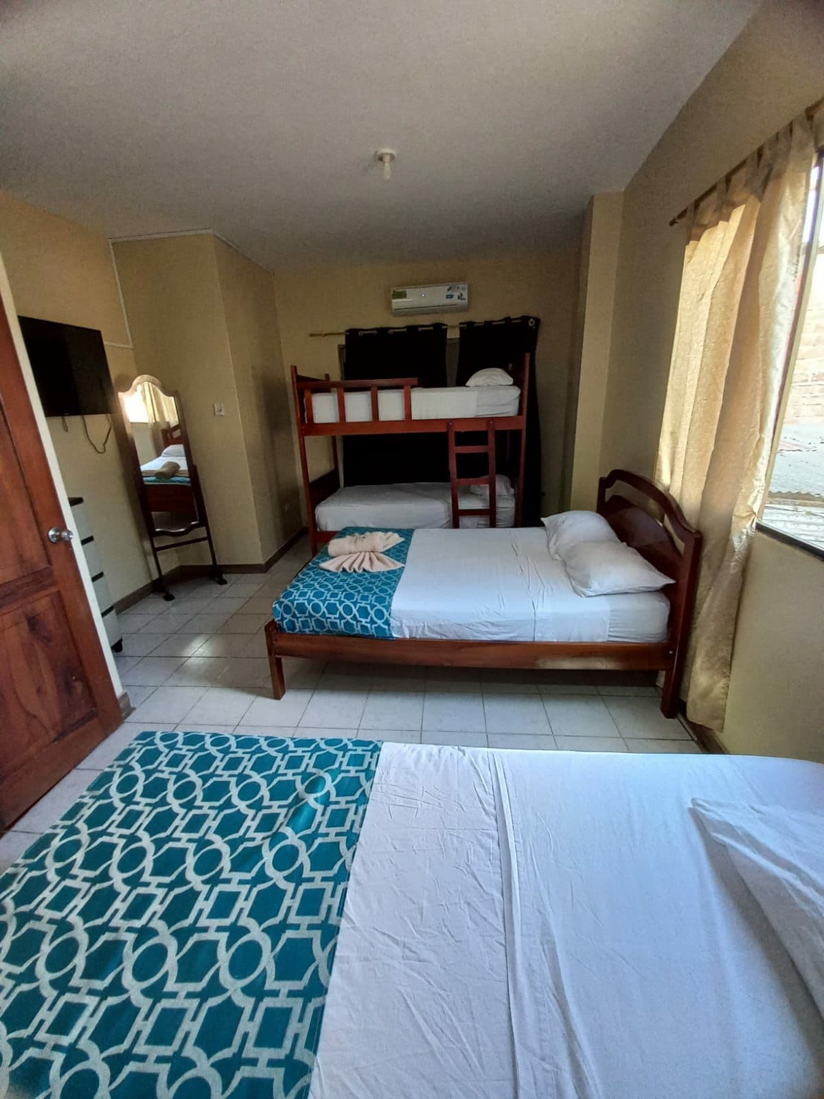 A shared sleeping area is equipped with a double bed and a bunk bed. Light-colored walls enhance the brightness from natural light filtering through window curtains. A decorative rug adds texture to the space, while a mirror and a wall-mounted television are positioned nearby.