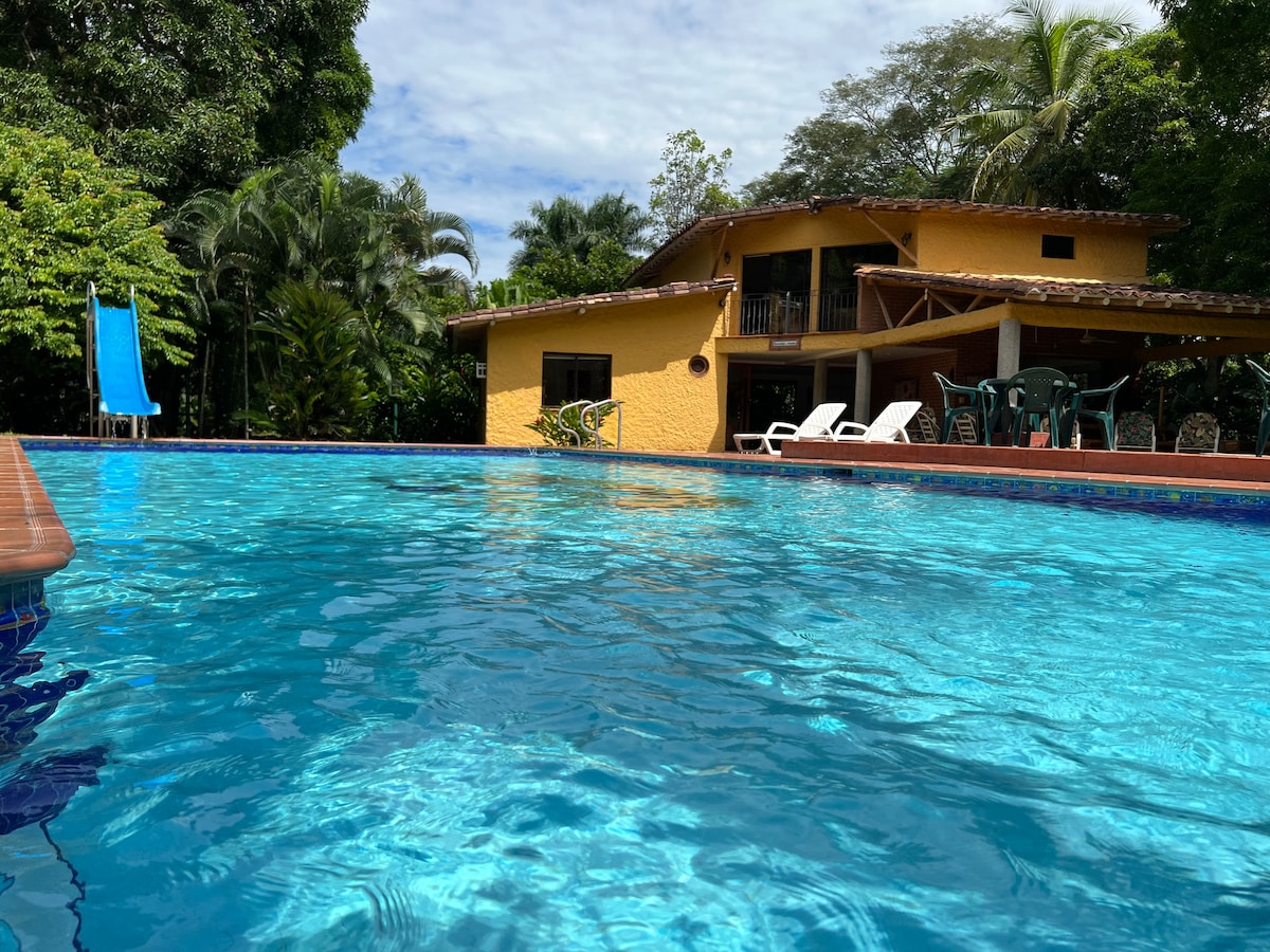 A clear view of a large pool reflects the vibrant blue sky, surrounded by lush greenery. The two-story yellow finca is visible in the background, featuring a balcony and outdoor seating areas. A blue lounge chair and white sunbeds are positioned near the pool's edge.