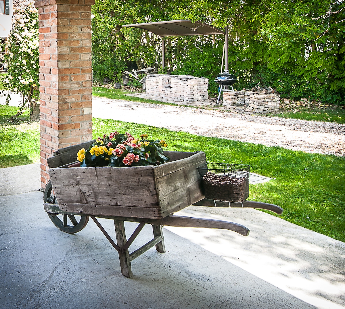 A rustic wooden wheelbarrow filled with vibrant flowers is positioned on a concrete surface. In the background, a shaded area features a grilling station and additional brick structures, surrounded by lush greenery and a well-maintained lawn.