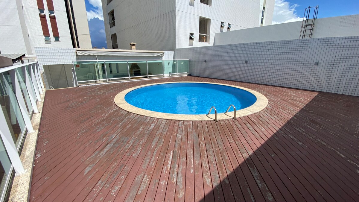 A circular pool is set within a wooden deck, surrounded by a white tiled wall. Natural light reflects off the water's surface, while nearby glass panels enhance the openness of the space. The area appears well-maintained and spacious.