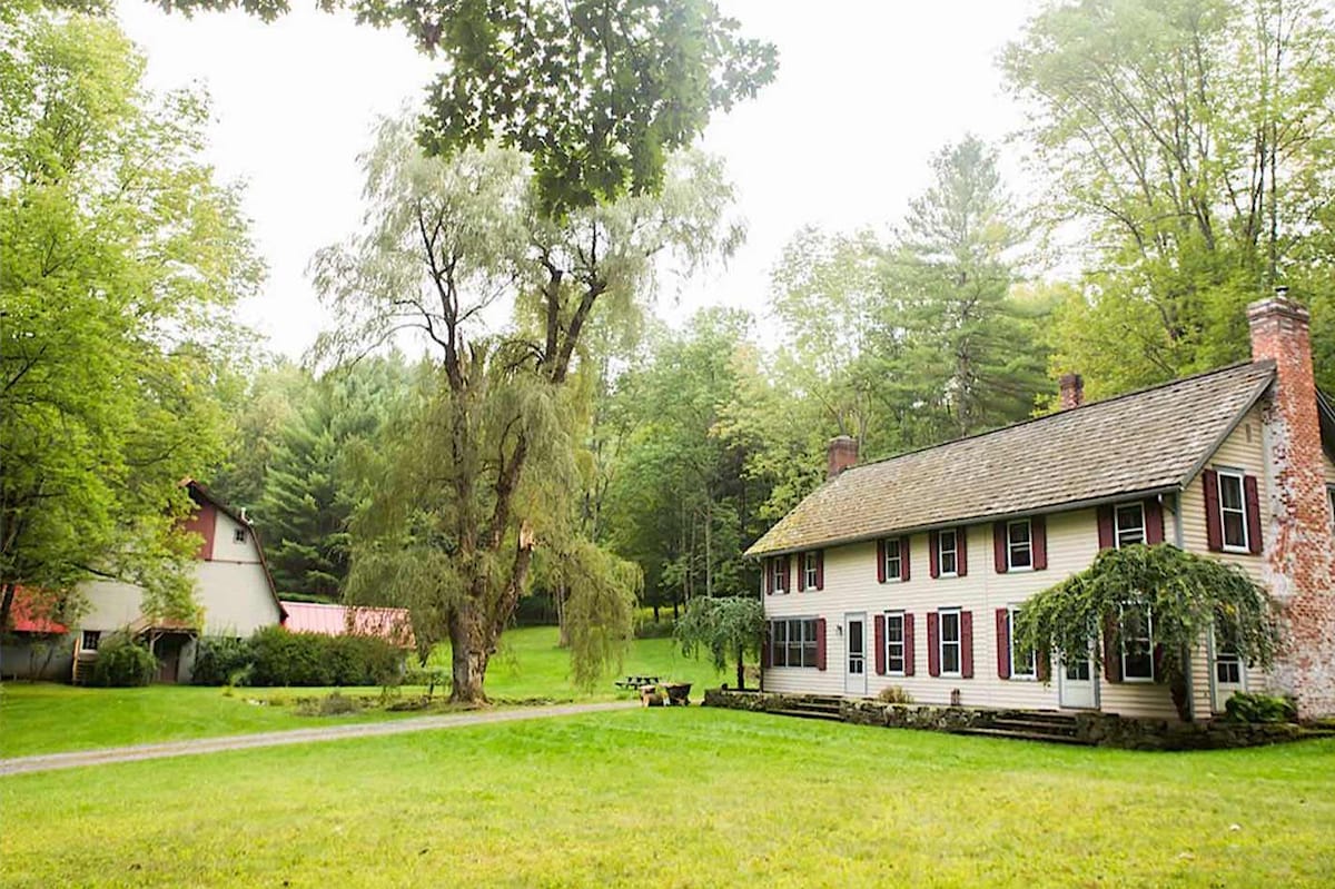 A charming two-story house is set among lush greenery, with large windows framed by red shutters. Adjacent to the house, a rustic outbuilding can be seen, both surrounded by open grassy areas and trees, creating a serene outdoor setting.