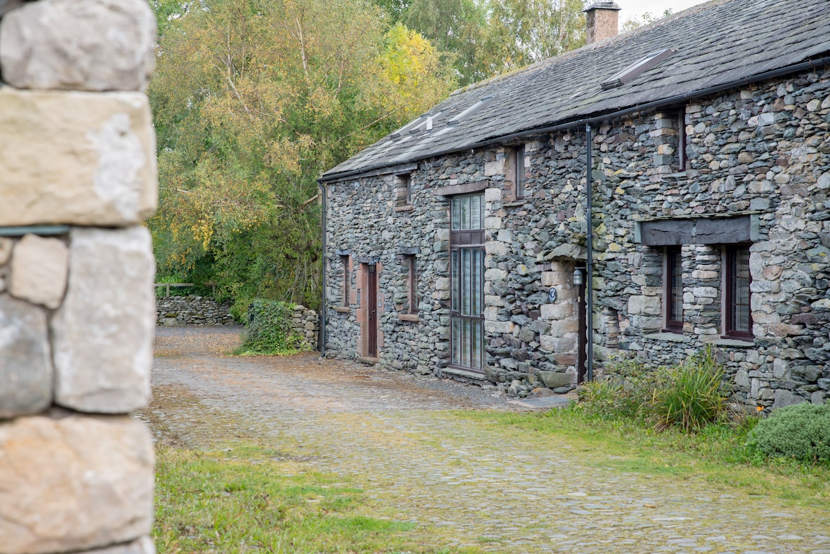 A stone building with a rustic facade is shown, featuring several windows and doors. The pathway leading to the entrance is bordered by greenery and trees, enhancing the natural setting of the property.