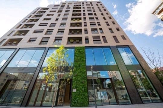 The modern building stands tall with a facade of large glass windows, reflecting the surrounding area. A green vertical garden is integrated into the design, adding a touch of nature to the contemporary architecture. The sky above features soft clouds, enhancing the overall ambiance.
