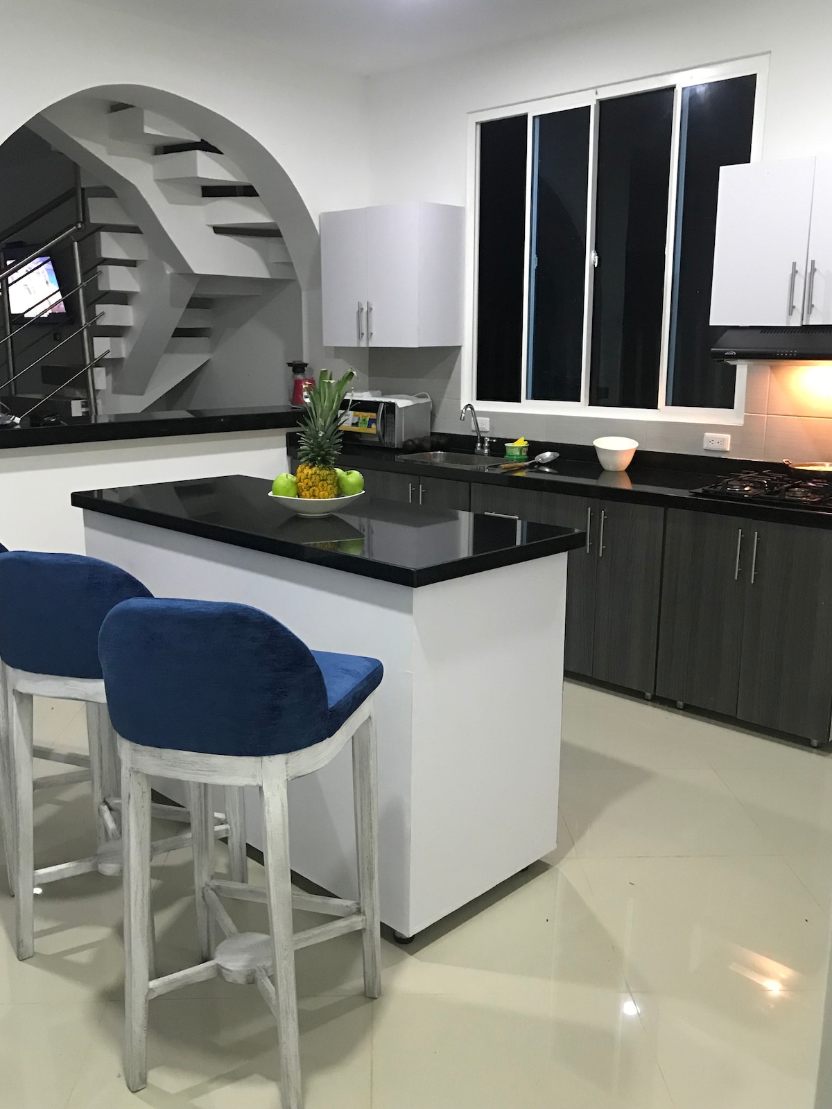 A modern kitchen is presented with sleek black countertops and light cabinetry. Two bar stools feature soft blue upholstery, positioned at a central island. Large windows allow natural light to illuminate the space, highlighting the contemporary appliances and the minimalist design.