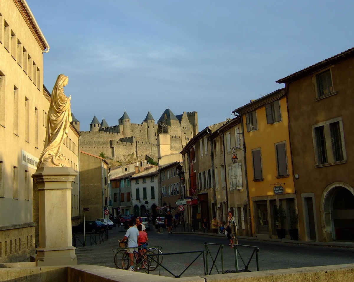 A charming village street is captured, featuring colorful buildings and a statue on the left. A couple rides bicycles while others stroll. In the background, a historic castle stands tall under a clear sky, enhancing the village's scenic appeal.