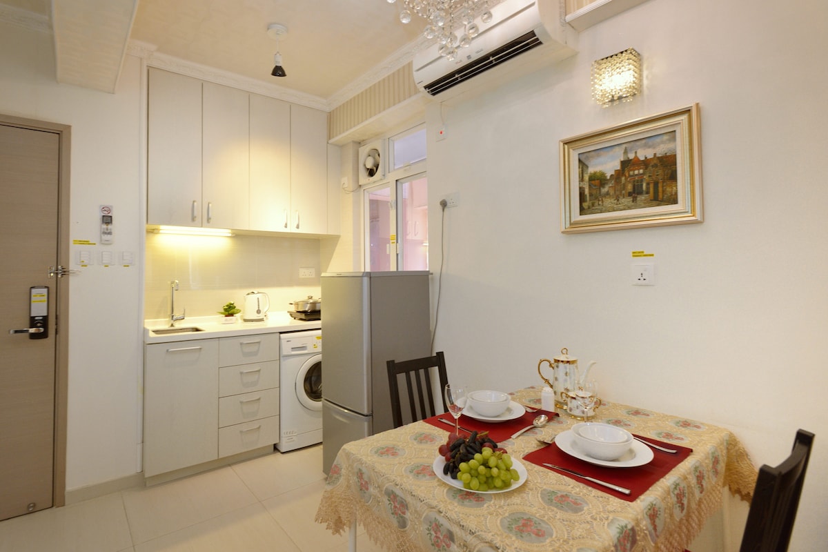 A dining area features a table set for two, complete with plates and a teapot. The kitchenette, equipped with a refrigerator and washing machine, is visible in the background. Light is provided by a stylish ceiling fixture, enhancing the clean and modern design.