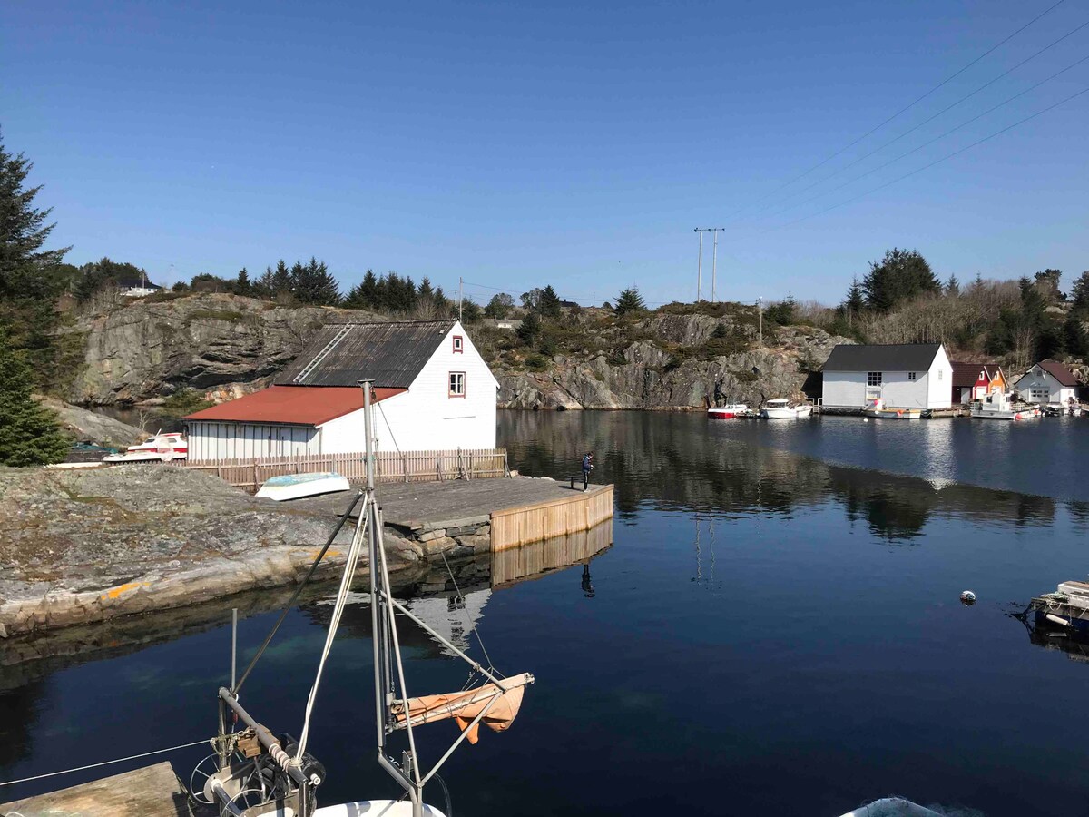 A waterway reflects the clear sky, bordered by rocky formations and quaint buildings. A sailboat rests at a wooden dock. Several boats are anchored in the calm water, while trees and landscapes frame the scene, contributing to a peaceful coastal environment.