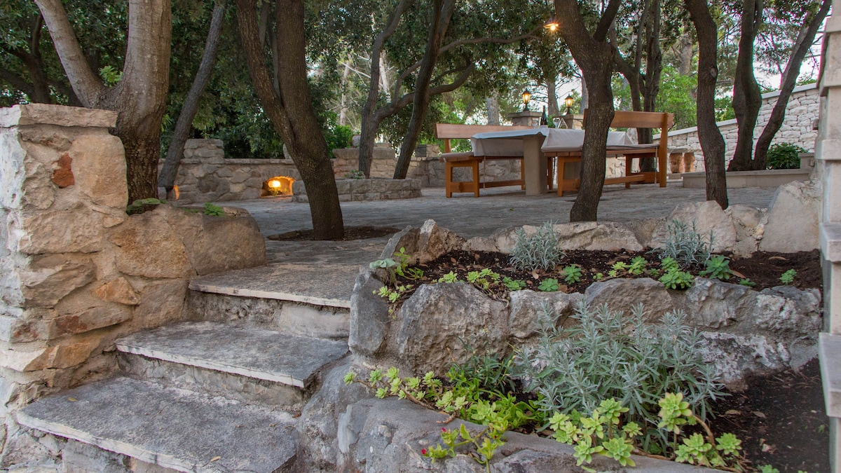 A stone pathway leads past lush greenery and a small garden, guiding to a dining area. A wooden table and benches are sheltered under the natural shade of oak trees, creating a serene space for outdoor meals.