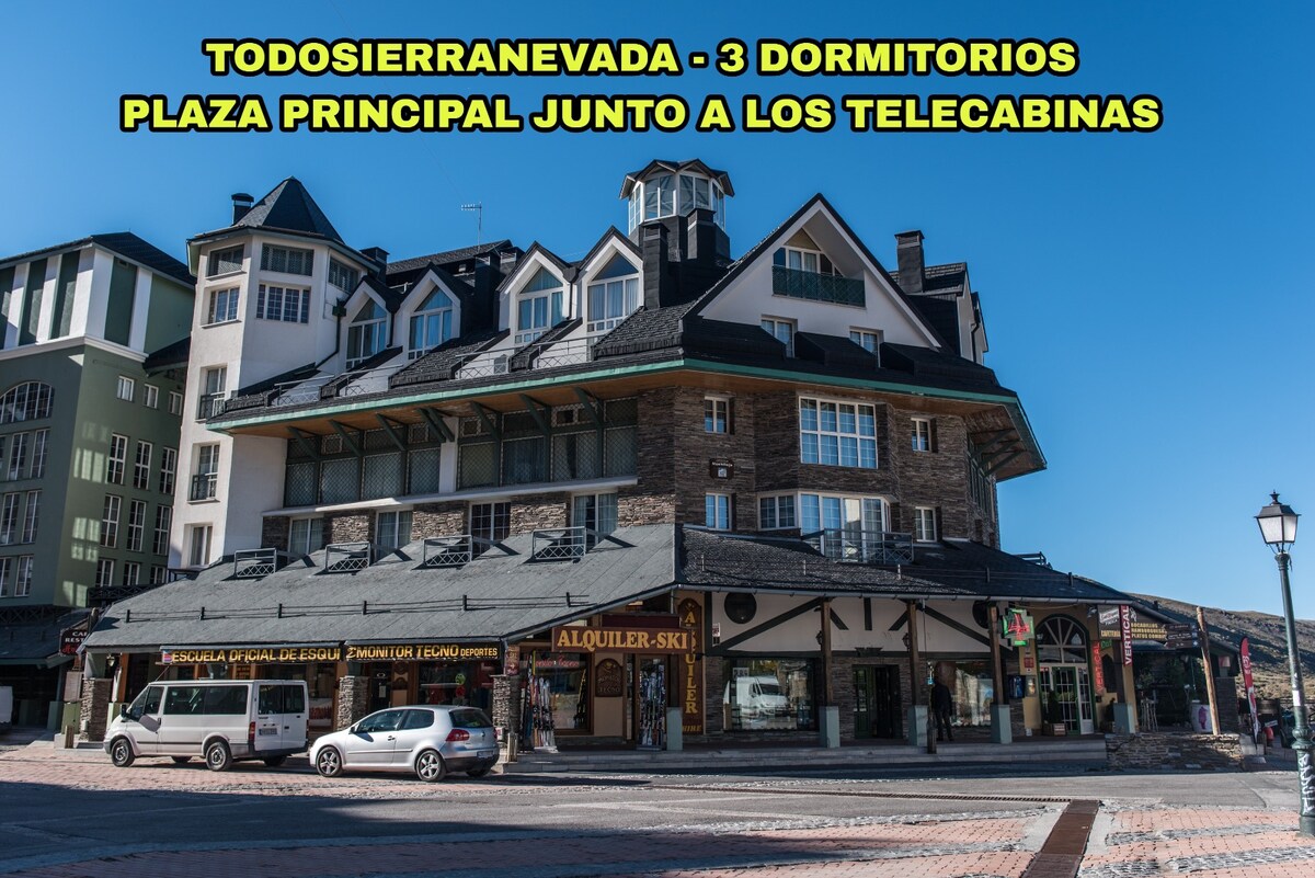 The image features a multi-story building situated in a lively plaza, directly beside the ski lift. The structure showcases a mix of stone and wood elements, with numerous windows allowing natural light. Signs for ski rentals and nearby restaurants are visible on the ground level.