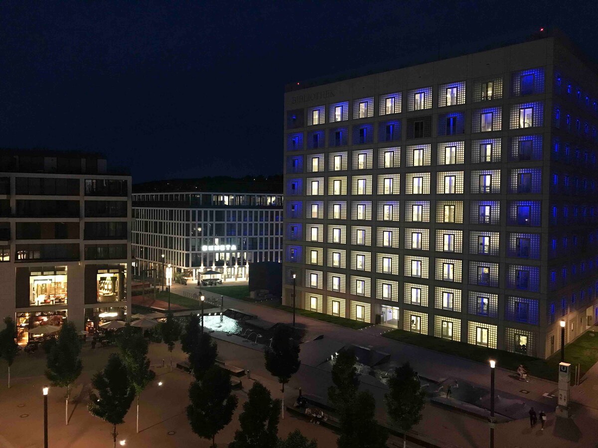 A modern architectural complex is featured at night, showcasing vibrant blue lighting on one building's facade. The surrounding area includes landscaped trees and an open plaza that invites exploration, while large windows across the buildings reveal warm interior lighting.