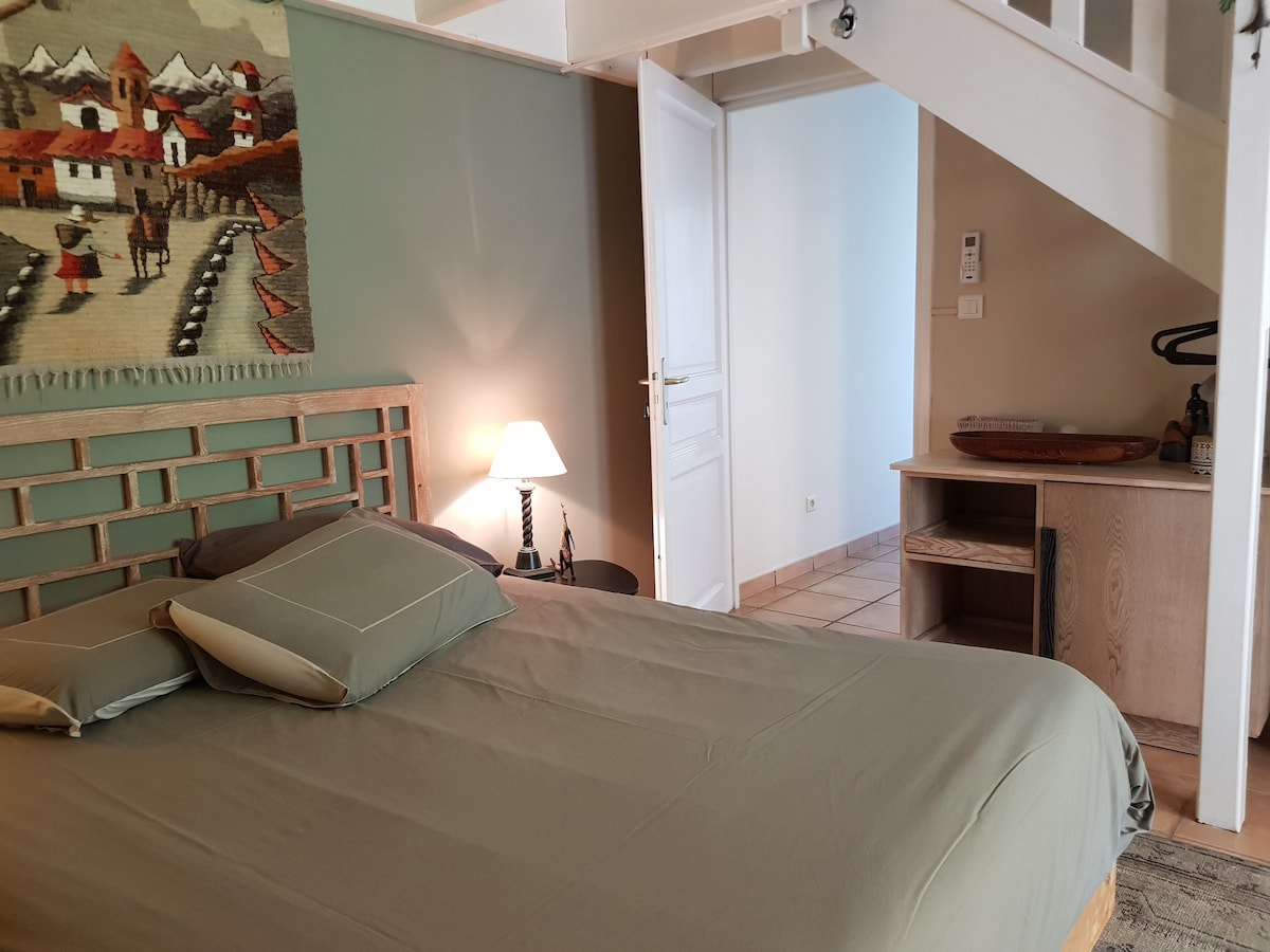 A spacious bedroom is depicted, featuring a large bed with soft linens and accent pillows. A bedside lamp provides gentle lighting, while a decorative wall hanging adds color. An open door leads to an adjoining space, and a simple wooden dresser is visible nearby.