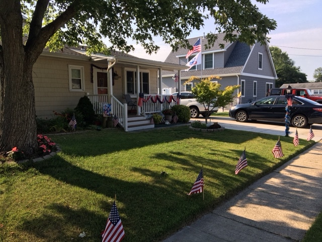The exterior of the rental unit is presented, featuring a welcoming porch adorned with American flags and red, white, and blue bunting. A well-maintained lawn, bordered by small garden beds, showcases a variety of flowers, while a driveway accommodates one vehicle.
