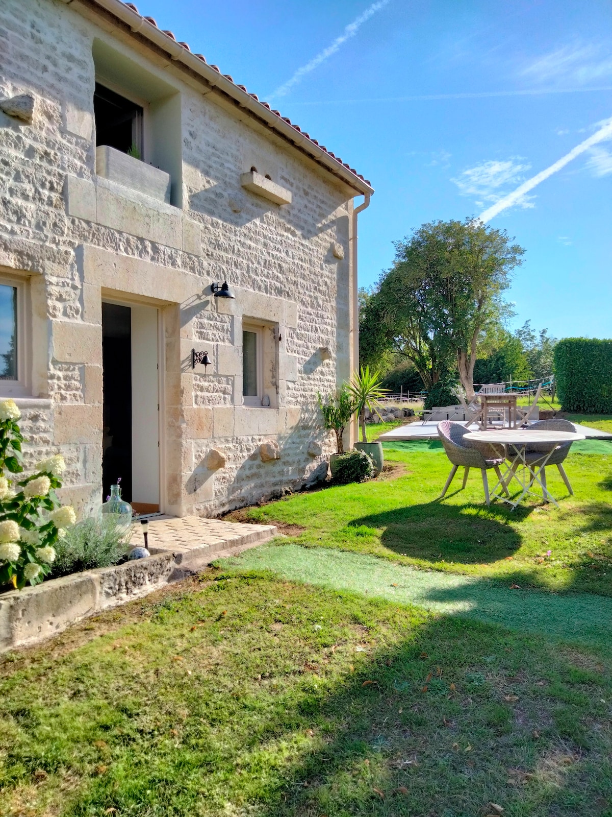 The exterior of the stone house is highlighted by sunlight, showcasing a welcoming entrance. A grassy area features a wooden dining table surrounded by chairs, set against a backdrop of trees and landscaped greenery.