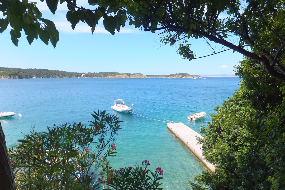 The image captures a serene waterfront view featuring two boats, gently bobbing in the clear blue water. Lush greenery frames the scene, while a stone jetty extends into the water, providing access to the serene surroundings. Hills can be seen in the background, enhancing the natural landscape.
