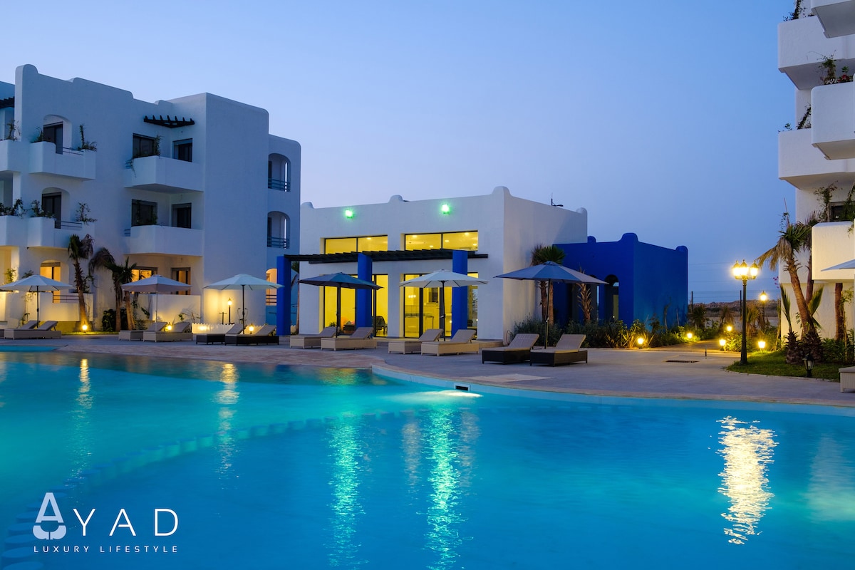 The image shows a serene swimming pool with illuminated blue water, inviting relaxation. Modern buildings with balconies are visible in the background, and outdoor seating areas with umbrellas are arranged around the pool, creating an atmosphere suited for leisure and enjoyment.