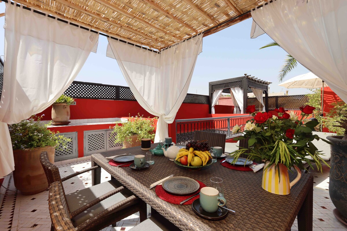 A shaded terrace features a dining table surrounded by chairs, with decorative plants and flowers arranged around the space. Plates and cups are set on the table, alongside a bowl of fresh fruit. Light drapes gently hang to create a cozy atmosphere.