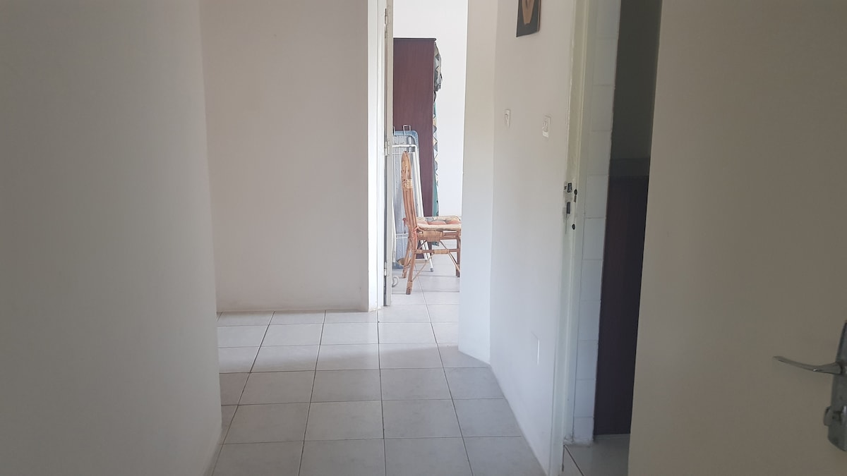 An unobtrusive hallway is displayed, featuring light-colored walls and tile flooring. Natural light enters through an open doorway, illuminating a wooden rocking chair positioned in the background. The space conveys an open and inviting passage to the adjoining rooms.