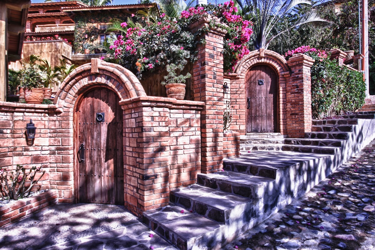 A charming entrance features two wooden doors set within arched brick frames, surrounded by vibrant flowering plants. Sturdy stone steps lead to the doors, casting intricate shadows on the textured pathway below.