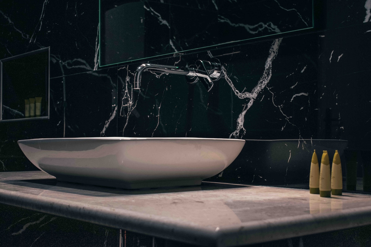 A sleek sink with a contemporary design is positioned on a marble countertop. Dark marble walls provide a striking backdrop, while minimalistic decor complements the modern aesthetic. Small containers are neatly arranged on the edge of the counter, emphasizing organized elegance.