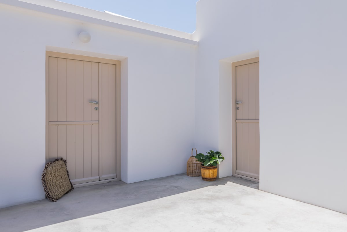 A serene outdoor area features two simple wooden doors with neutral-colored finishes. A small potted plant adds a touch of greenery beside a woven basket, while the smooth concrete floor reflects the bright sunlight.