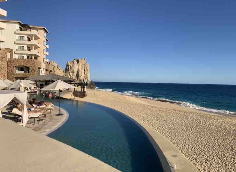 A tranquil pool stretches alongside a sandy beach, offering a seamless transition to the ocean. Umbrellas provide shade over lounge chairs, while large rock formations stand out against the clear blue sky. Waves gently lap at the shore, complementing the serene environment.