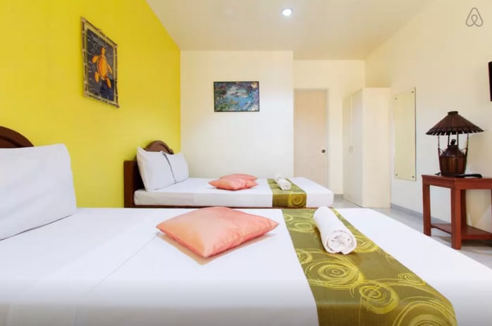 A well-lit room features two spacious beds dressed in crisp white linens, accented with orange throw pillows. A vibrant yellow wall adds color to the space. A wooden chair and table are positioned in one corner, while a small artwork adorns the wall.