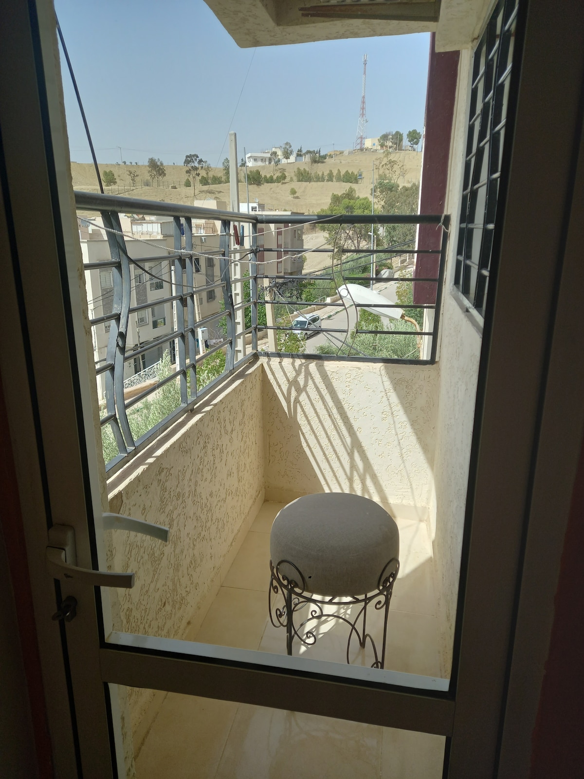 A small balcony is visible, featuring a round table with an ornate base. The unobstructed view includes distant hills and a broadcast tower. Natural light fills the space, with a simple railing providing security while allowing for outdoor enjoyment.
