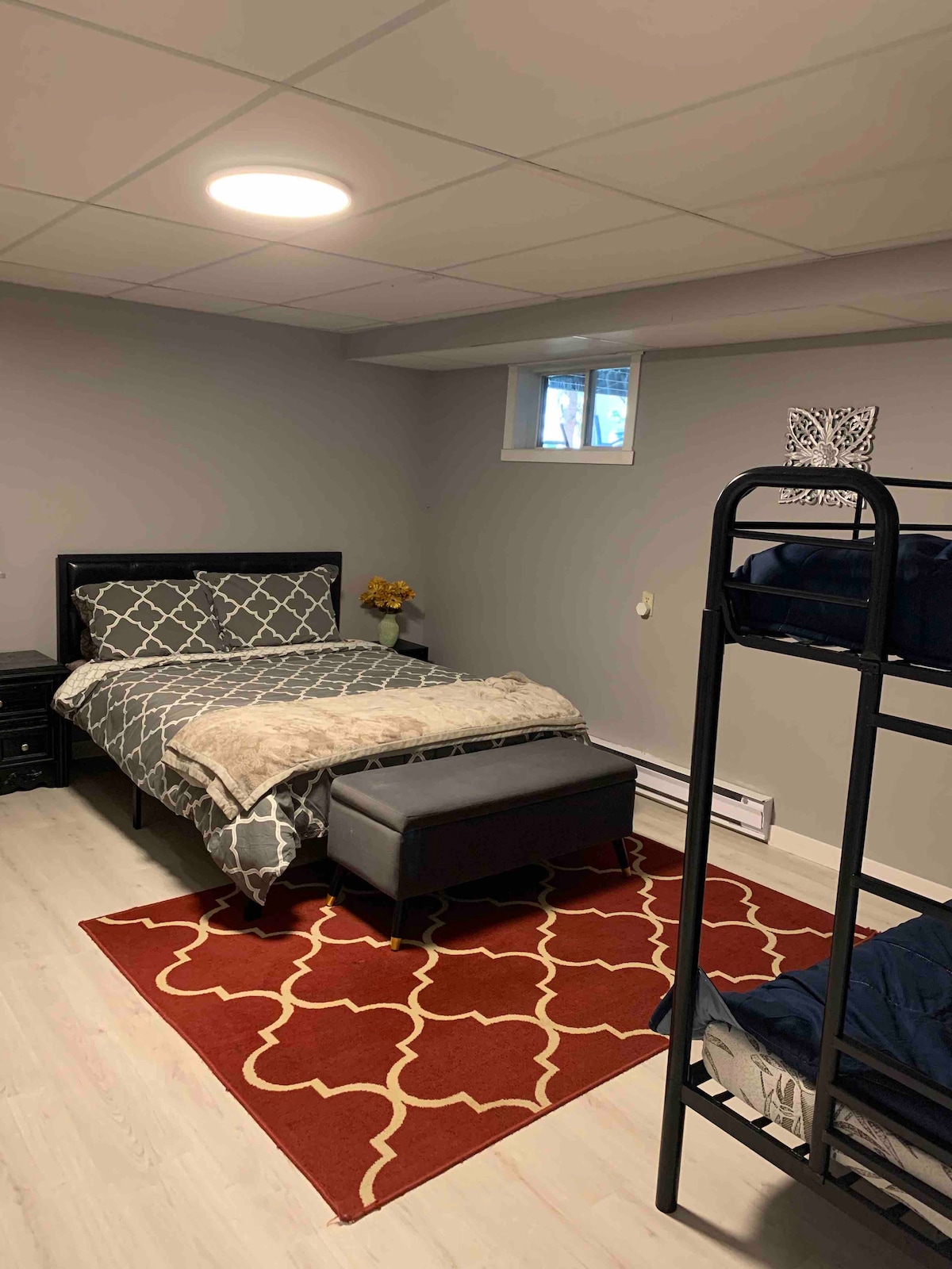 A spacious bedroom is presented, featuring a queen bed with a patterned blanket and a cozy ottoman at the foot. Bunk beds are positioned in one corner, and a vibrant area rug adds a pop of color to the light-colored flooring. A small window provides natural light.