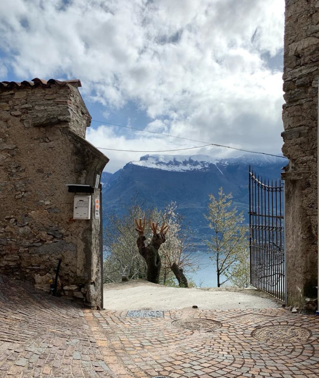 A stone pathway leads through a historic archway, showcasing a serene view of mountains lightly dusted with snow and a calm lake below. A large, bare tree stands nearby, with the sky filled with soft clouds providing a tranquil backdrop.