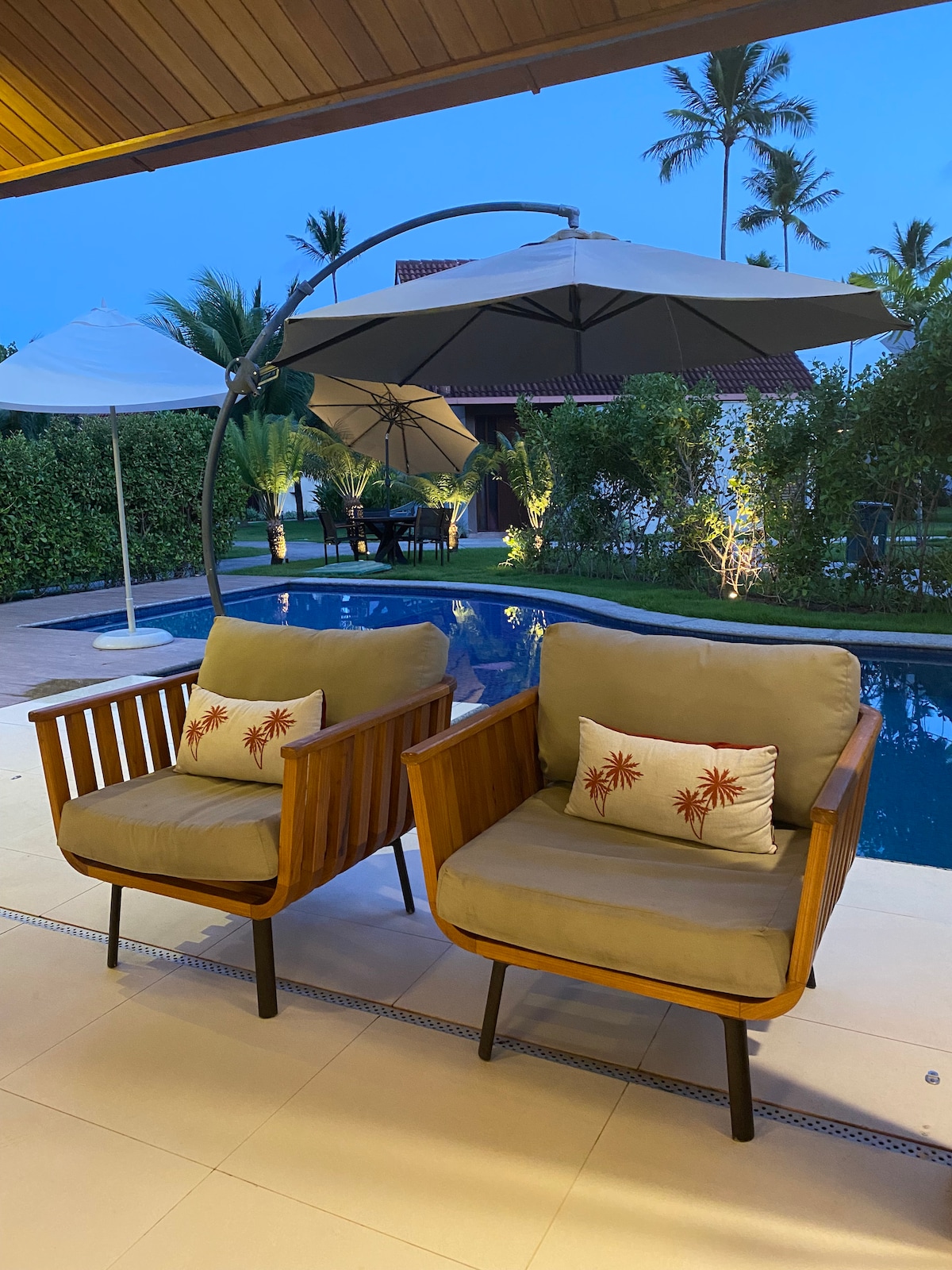 Two wooden armchairs with soft cushions are positioned near a private pool, surrounded by palm trees and parasols. Soft lighting highlights the tranquil area, creating a peaceful setting for relaxation.