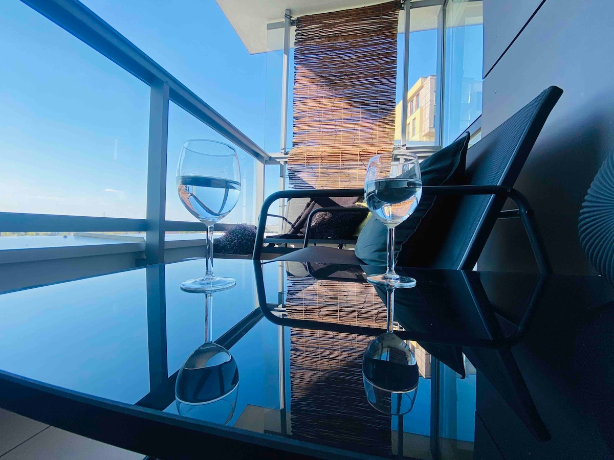 A balcony area is showcased, featuring a glass table reflecting the blue sky and nearby buildings. Two glasses of water are placed on the table, accompanied by a comfortable black chair and soft cushions, enhancing the scene with a touch of relaxation.