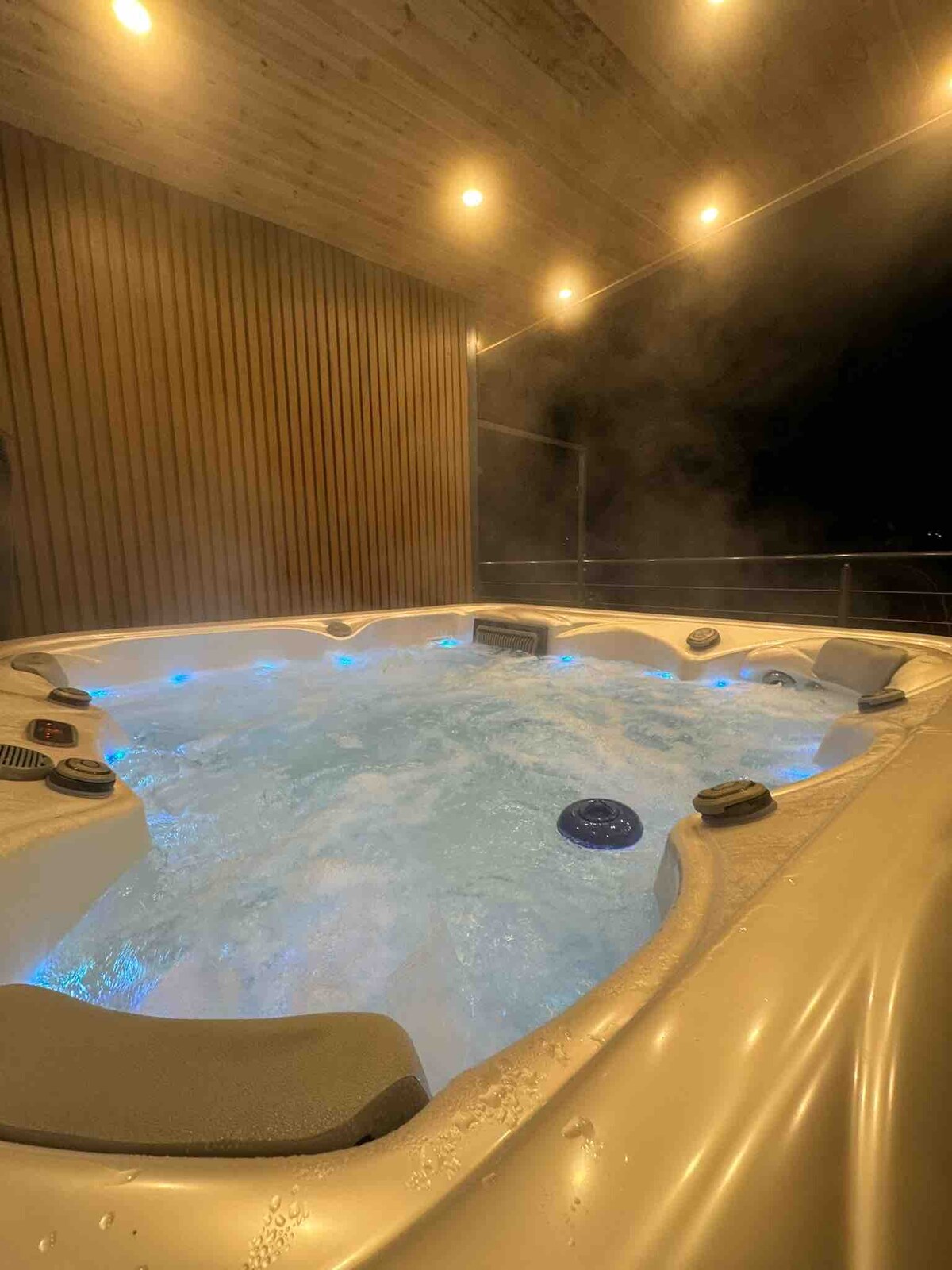 A private hot tub is illuminated by subtle blue lights, creating a serene atmosphere. Steam rises gently from the bubbling water, complementing the wooden paneling and overhead lighting that provide a soft glow, suggesting a relaxing retreat in a peaceful setting.