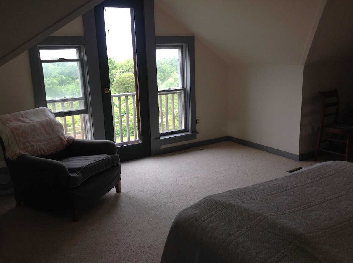 A spacious room is highlighted by large windows providing views of the surrounding greenery. A comfortable chair is positioned in one corner, complemented by soft carpeting underfoot. A cozy bed with a textured coverlet is visible, creating an inviting sleeping area.