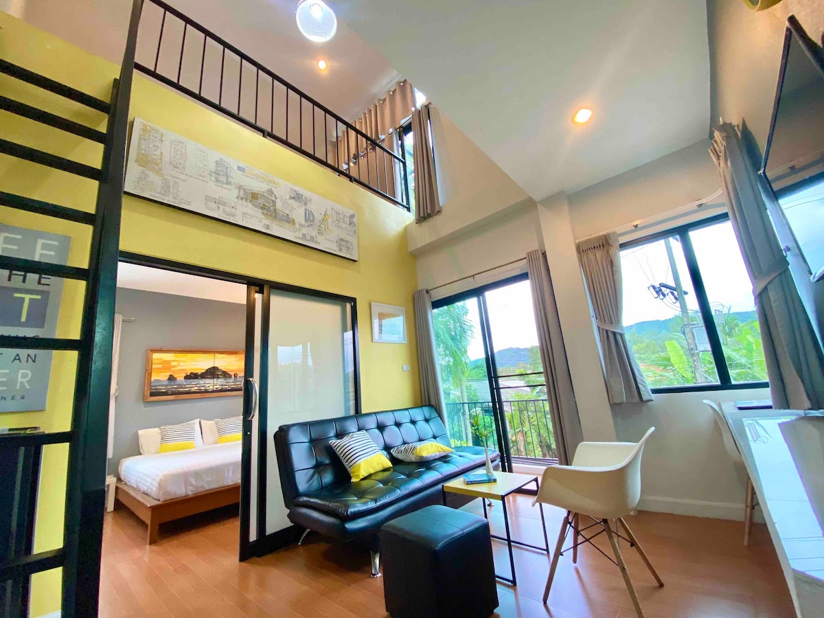 A modern living area features a sleek black sofa and minimalist decor. Large windows allow natural light to fill the space, showcasing views of greenery. A cozy sleeping area is visible through an open doorway, complemented by light wood flooring and neutral wall tones.