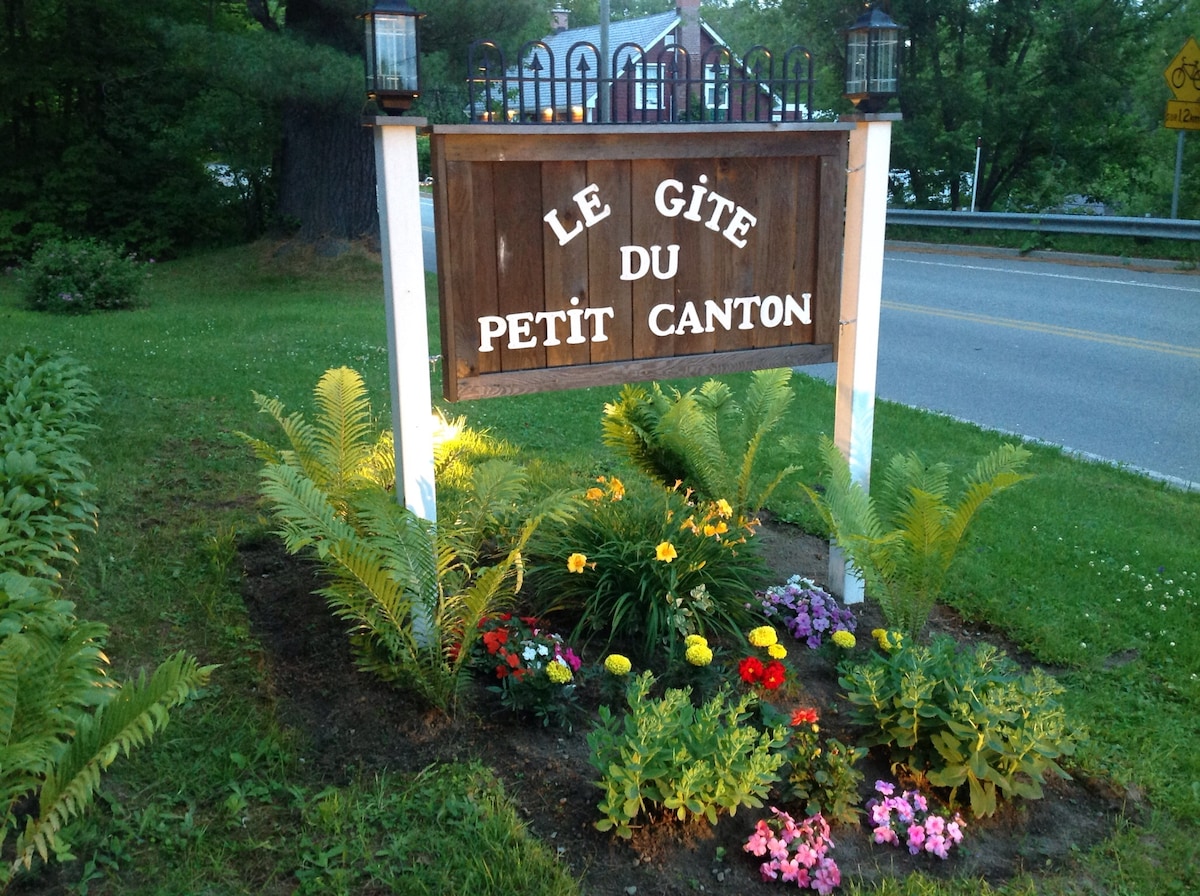 A wooden sign displaying 'Le Gîte du Petit Canton' is positioned prominently in a garden setting. Surrounding the sign are vibrant yellow flowers, various plants, and ferns, creating a welcoming entrance. This area is well-maintained and features a backdrop of trees and greenery.