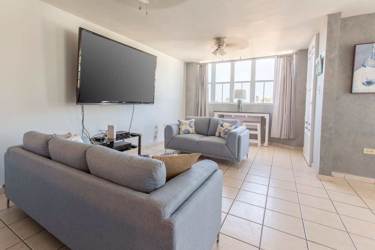 A light-filled living area features a large flat-screen TV mounted on a wall. Comfortable grey sofas are arranged to create an inviting seating arrangement. Large windows offer ample natural light, while neutral-colored walls and tiled floors contribute to the spacious atmosphere.