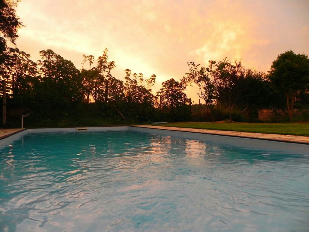 A serene swimming pool is visible under a colorful sunset sky, with hues of orange and pink reflecting on the water's surface. Lush greenery surrounds the area, contributing to a tranquil atmosphere.