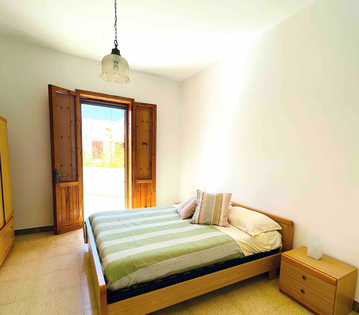 A bright sleeping area is illuminated by natural light from double doors leading to an outdoor space. A spacious bed with a striped green and white duvet is positioned against the wall. A wooden bedside table stands next to the bed, complementing the warm tones of the room.