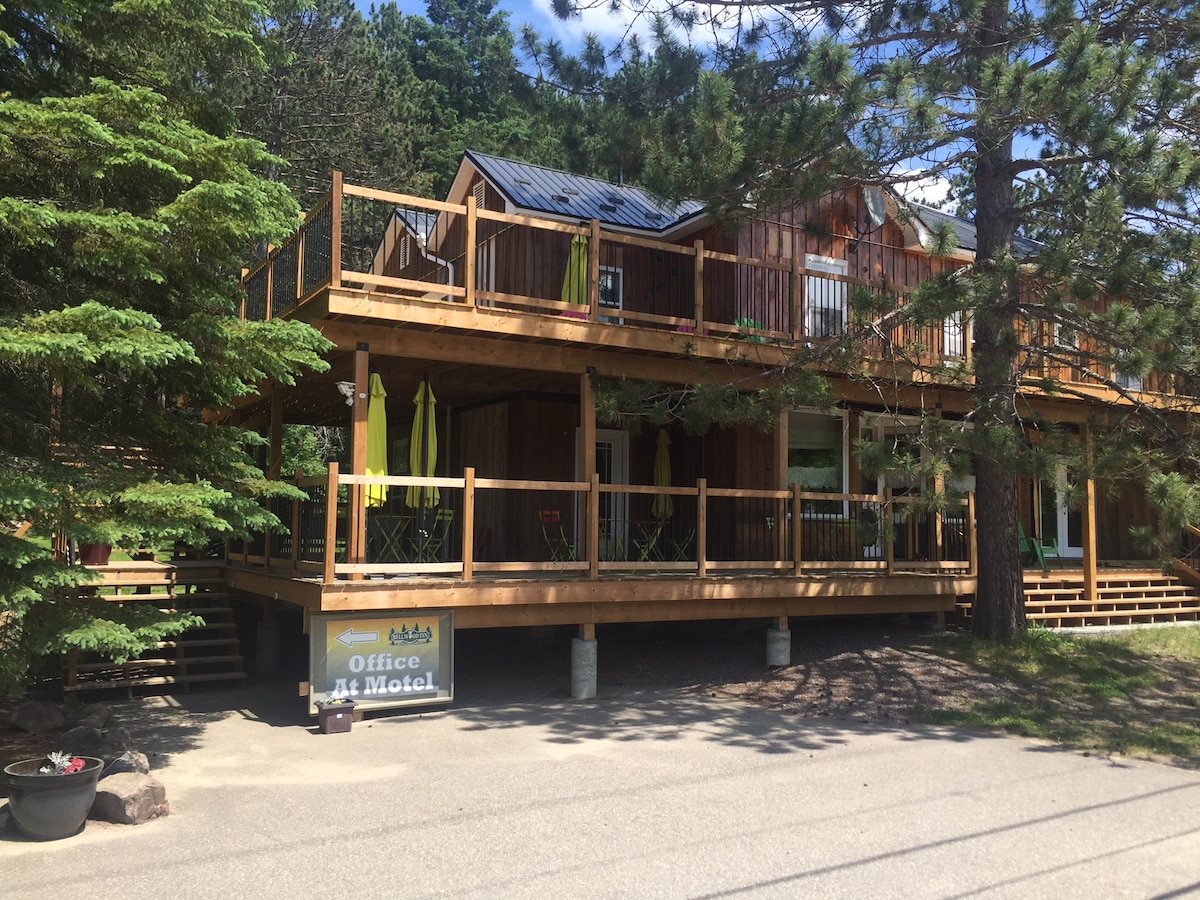 A wooden two-story structure features a spacious wrap-around deck, furnished with green and yellow chairs. A sign indicates the office entrance, surrounded by trees and a gravel path. The exterior showcases a natural wood finish, blending harmoniously with the outdoor setting.