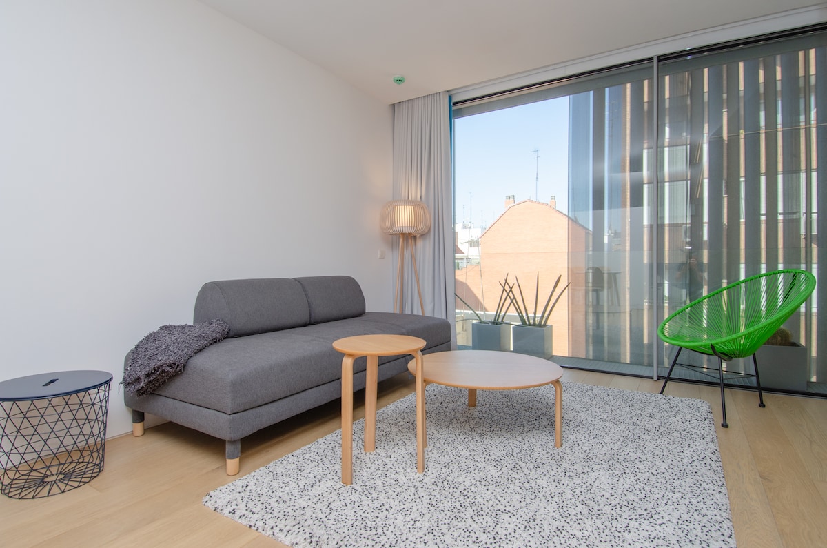 A modern living area is presented, featuring a gray sofa accompanied by two tables in different sizes. A woven rug lies beneath, and large windows create a bright atmosphere, displaying a view of the outside. An accent chair and potted plants add interest to the space.