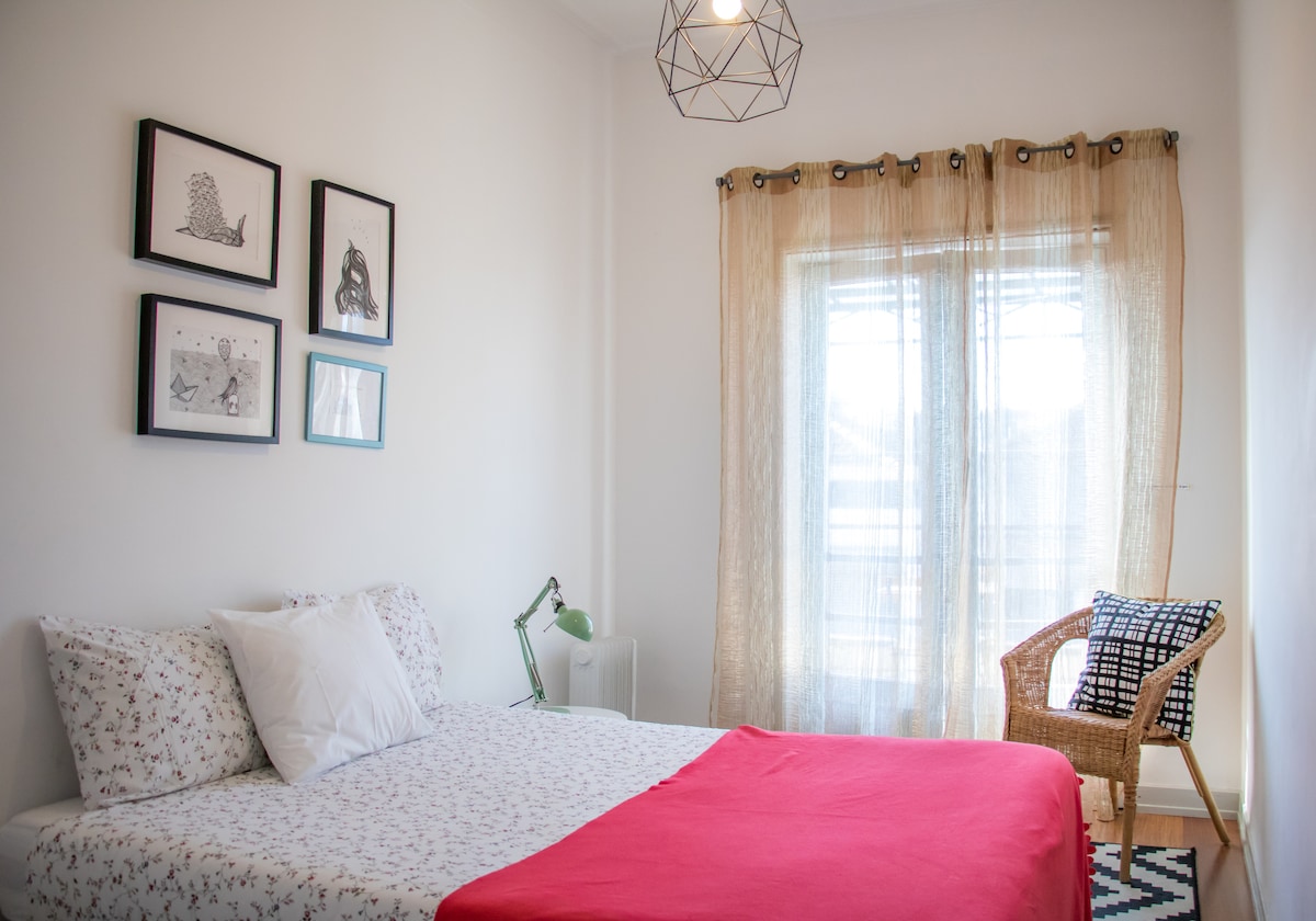 A bright and airy double room features a spacious bed with a patterned coverlet. A large window is framed by sheer curtains, allowing natural light to fill the space. Two framed artworks hang on the wall, and a wicker chair complements the decor.