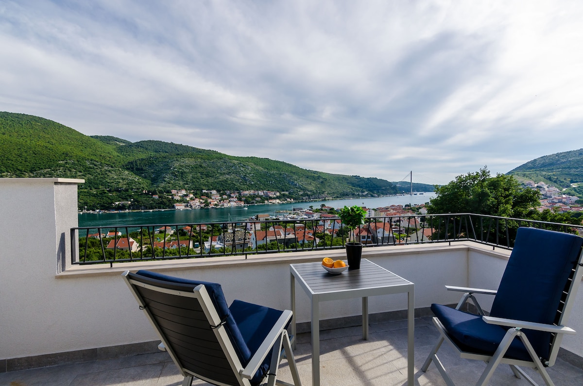 A peaceful outdoor terrace features two lounge chairs and a small table, overlooking the Ombla River and Dubrovnik Bridge. Lush green hills and picturesque houses are visible in the distance, creating a serene backdrop for relaxation.