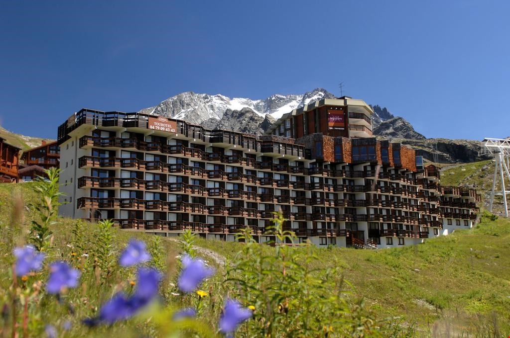 The exterior of the residence is showcased against a clear blue sky, with snow-capped mountains in the backdrop. A well-structured facade features multiple balconies and large windows, while vibrant wildflowers are visible in the foreground.