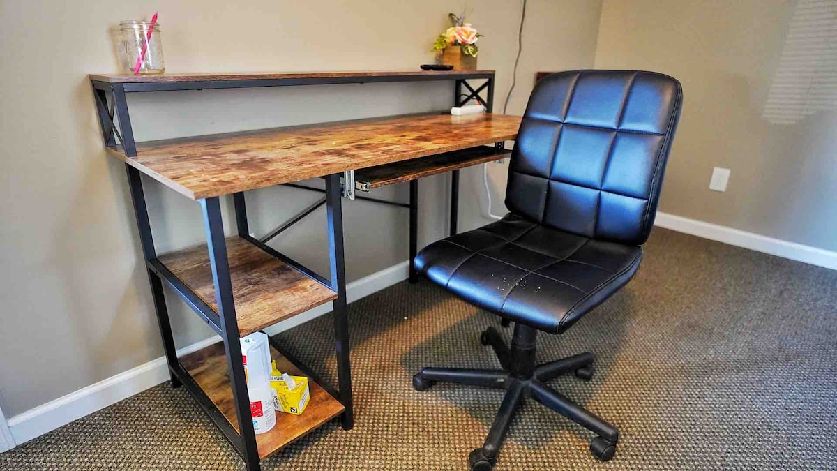 A simple workspace is featured, consisting of a wooden desk with a lower shelf for storage. A comfortable black office chair with a grid pattern sits in front of the desk. A small decorative plant adds a touch of color to the setup.